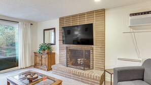 A living room with a fireplace, a flat-screen TV mounted above it, a sofa, a coffee table with decorative items, and a view through a sliding glass door.