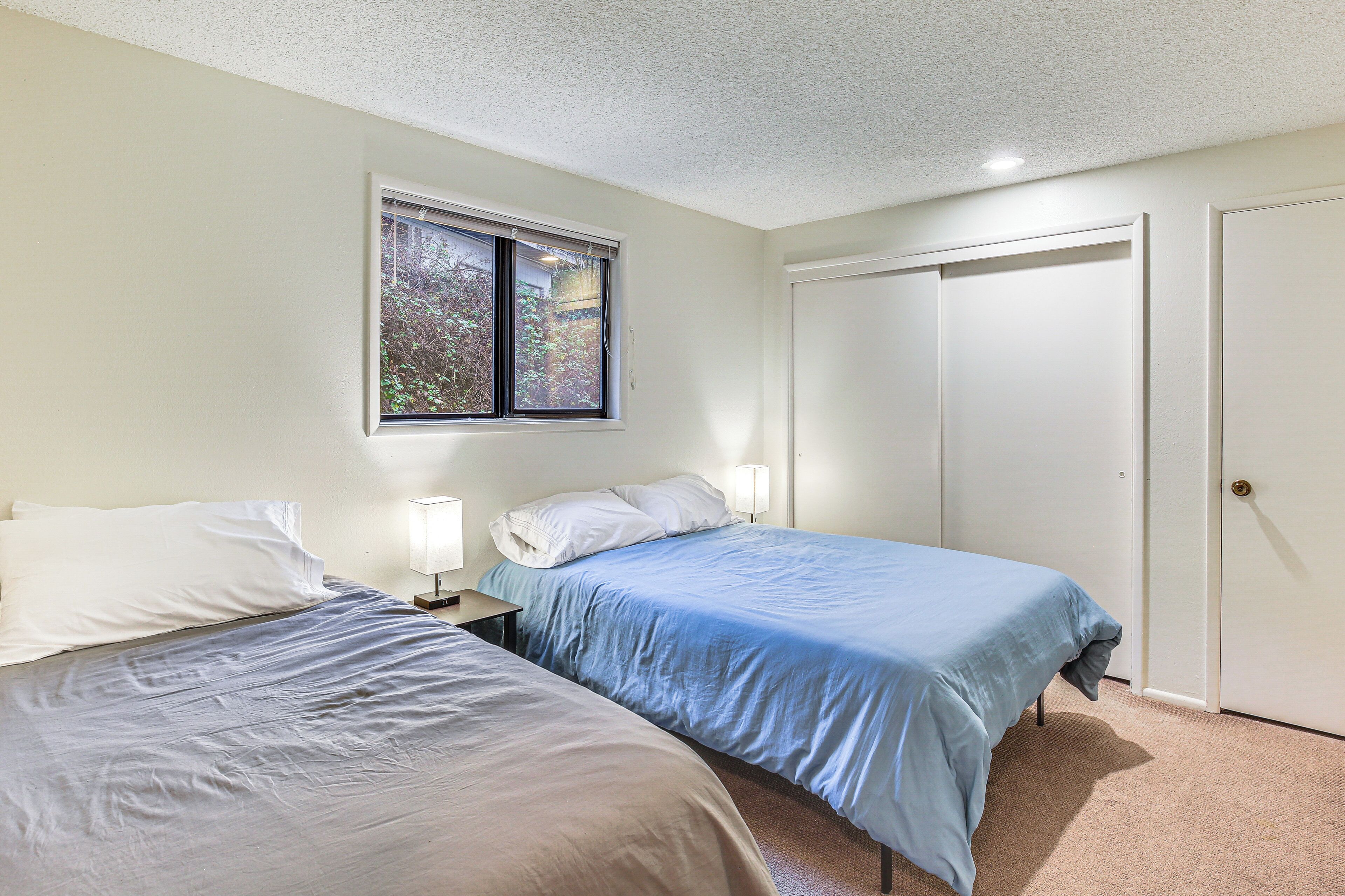 A bedroom with two beds, a window, a door, and a nightstand with a lamp.