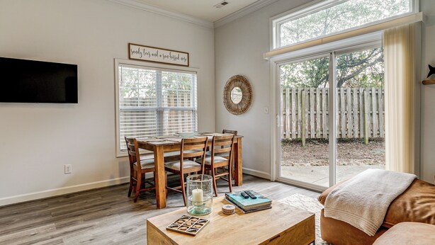 A dining area with a wooden table and chairs, a TV mounted on the wall, and a large window with a view of a wooden fence. - 4 Mi to Ocean: Modern Townhome in Wilmington! (Wilmington)