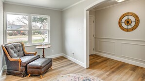 Una habitación con un ventanal grande, piso de madera, un sillón tapizado, una mesita redonda y un reloj de pared.