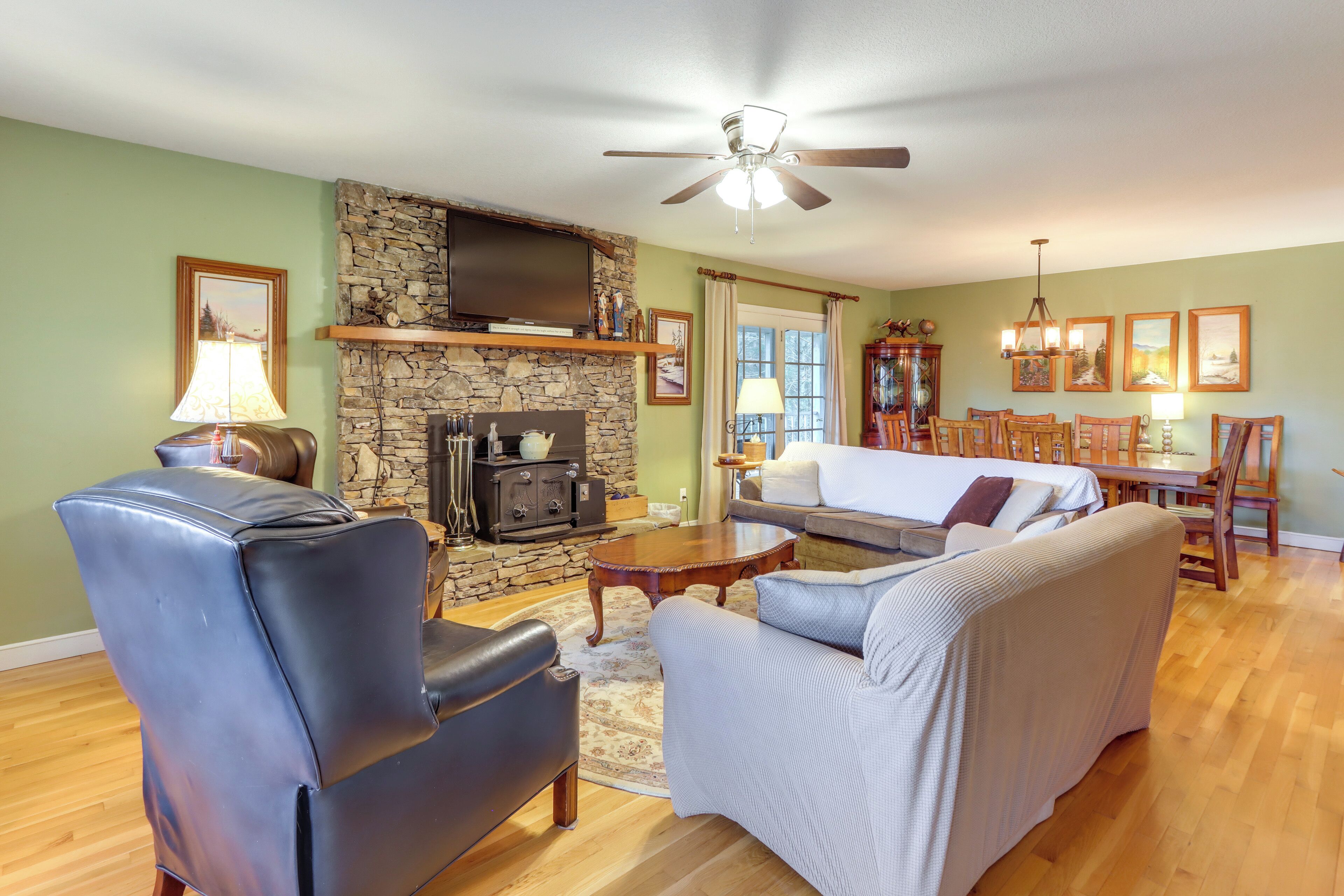 Sala de estar com lareira de pedra, poltronas de couro, sofá, piso de madeira e ventilador de teto.