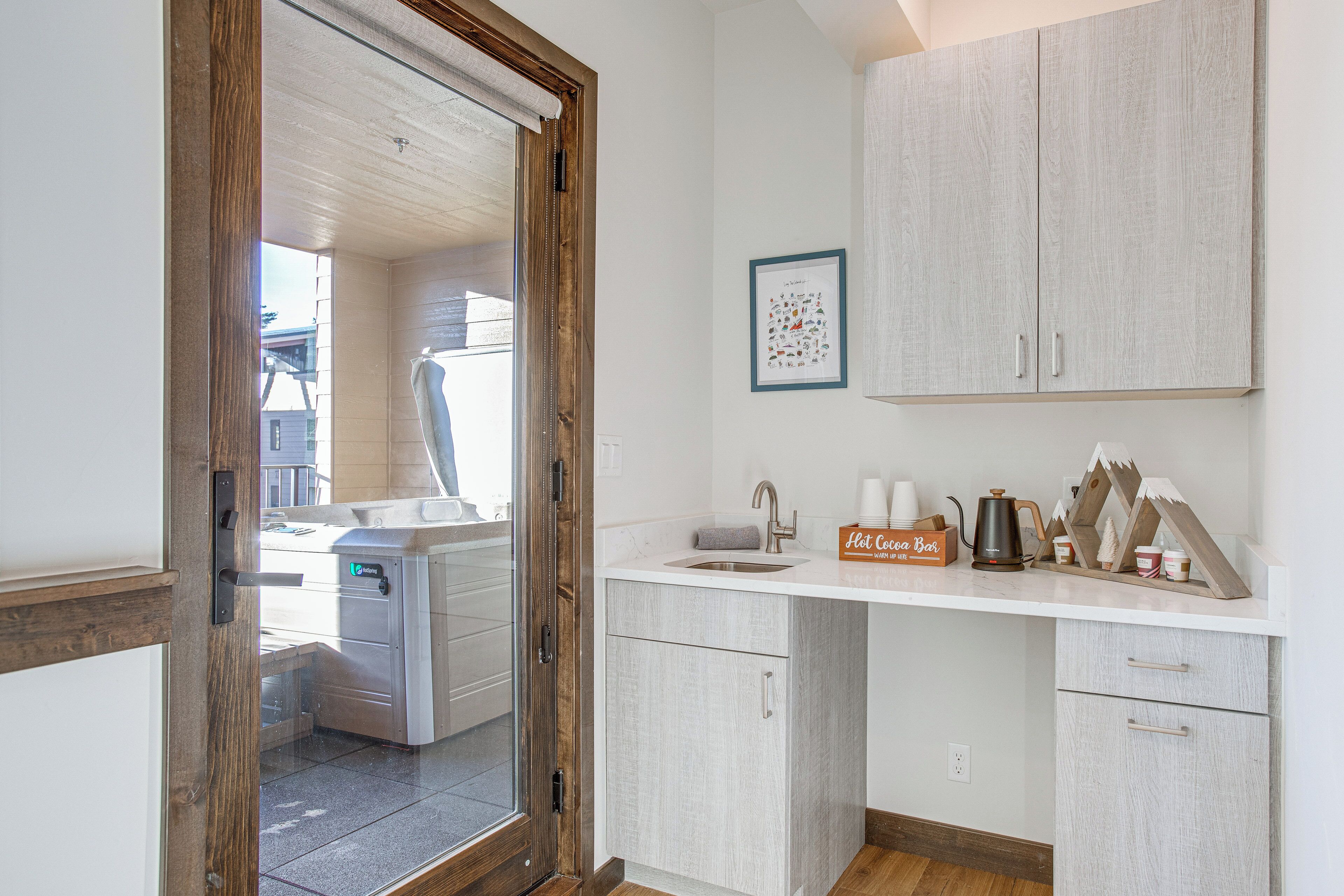 A modern kitchen with a wooden door, white cabinets, a sink, and a coffee station.