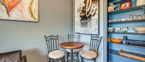 A dining area with a round table and four chairs, a bookshelf with various items, and two large abstract paintings on the wall.