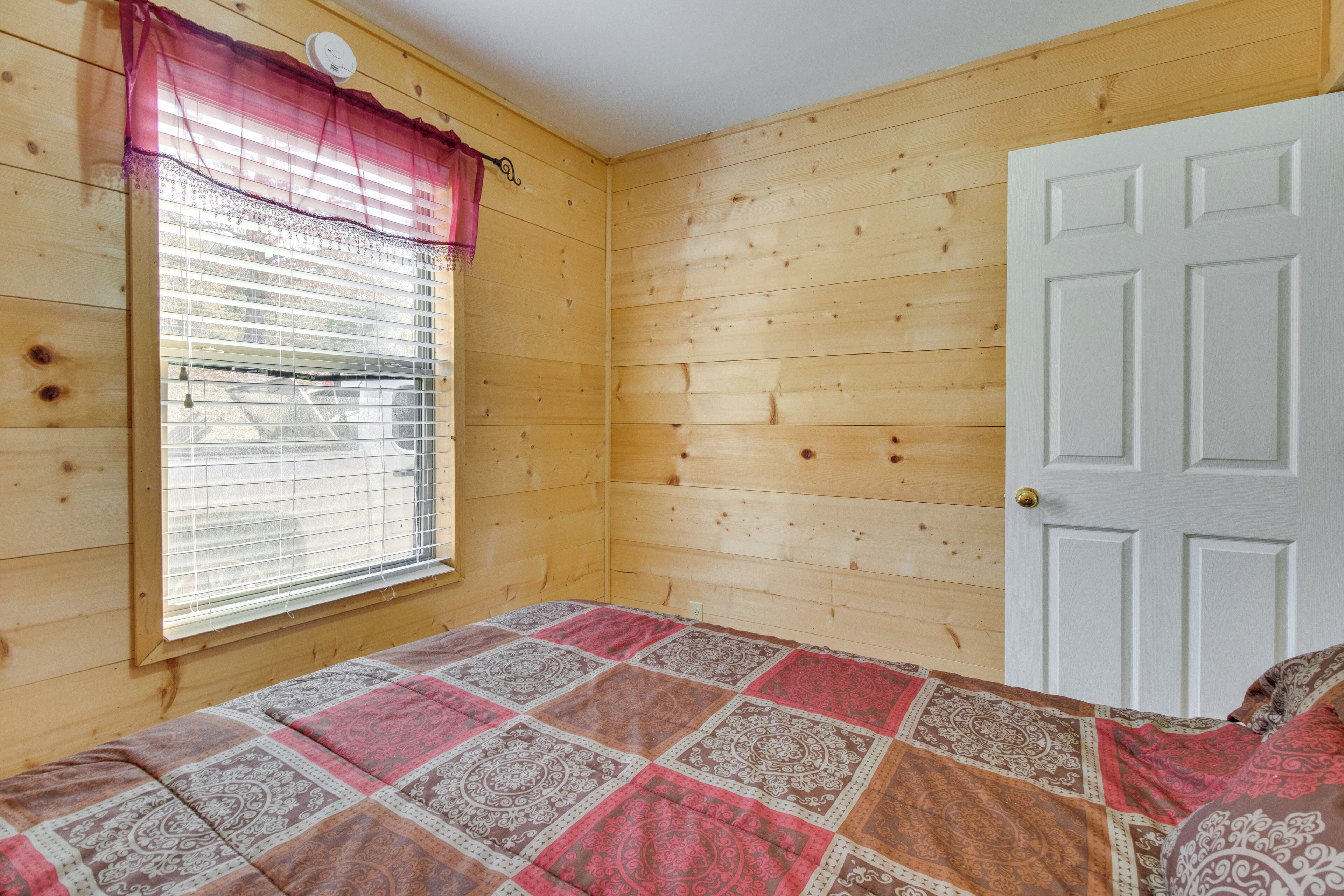 Una habitación de madera con una cama, un ventanal con persianas y una puerta blanca.