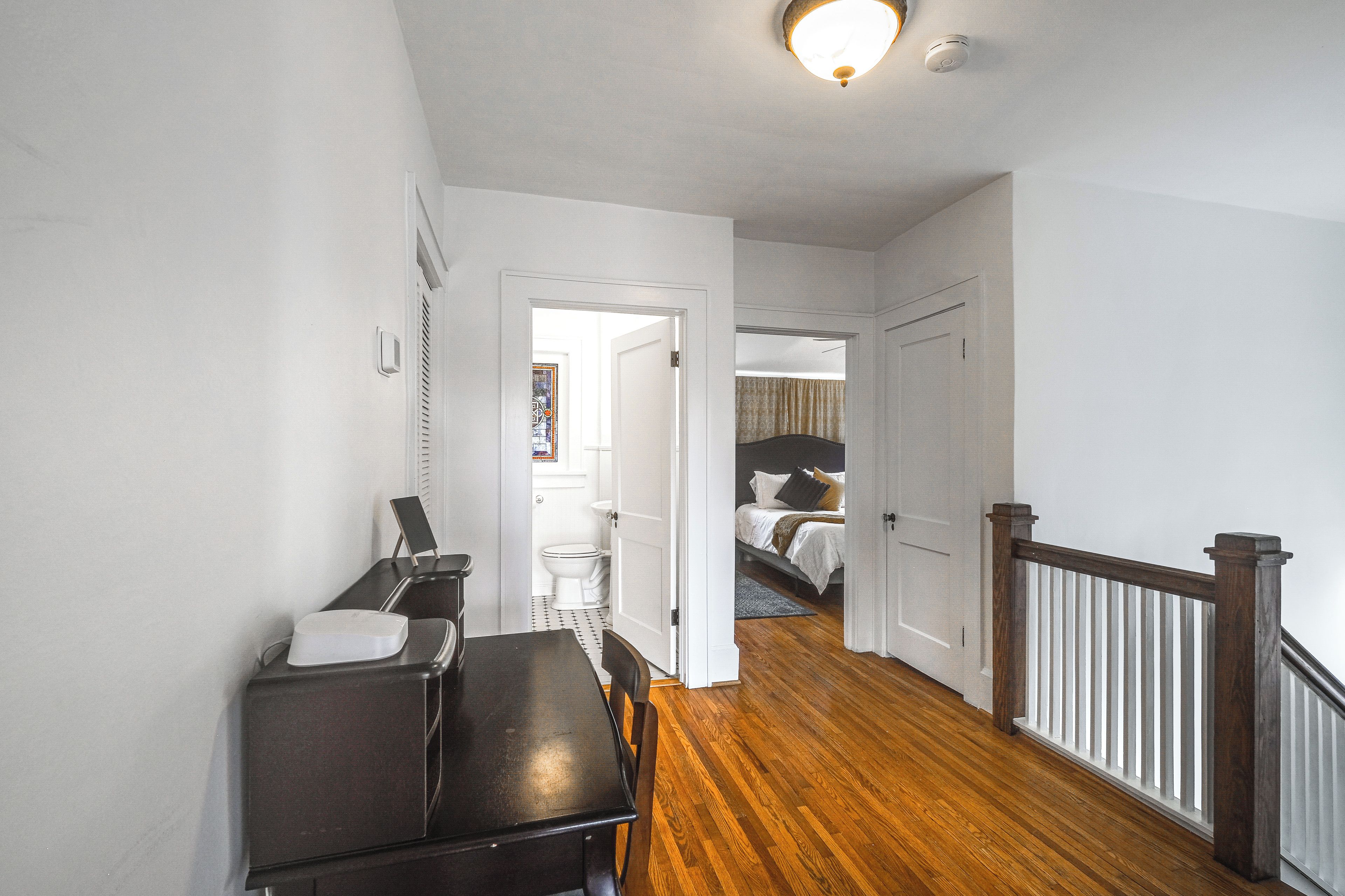 A hallway with a dining table and chairs, a staircase with a wooden railing, and a bedroom with a bed and a nightstand.