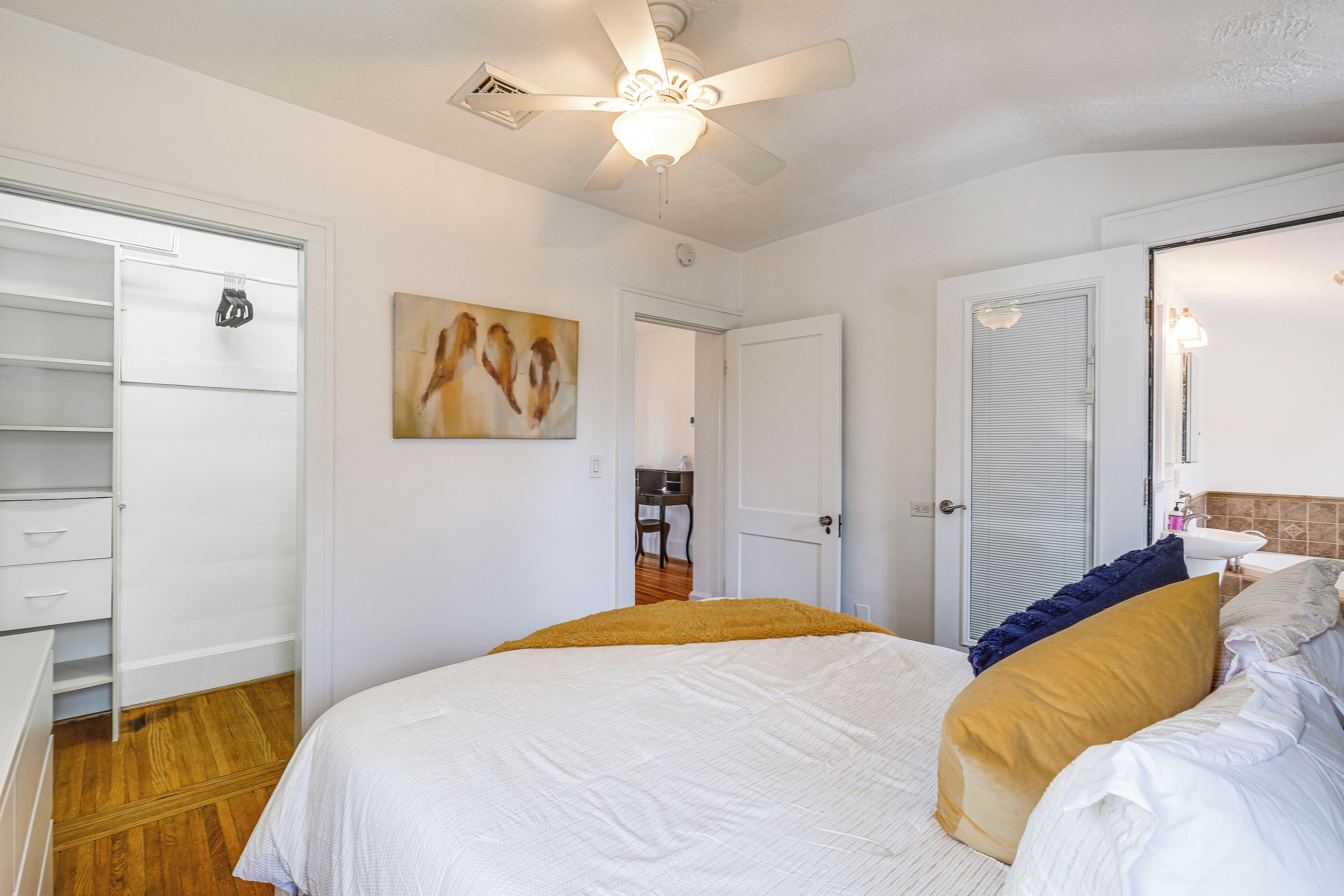 A bedroom with a bed, a ceiling fan, a painting, a wardrobe, and a bathroom with a sink and mirror.