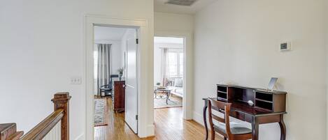 A spacious room with hardwood flooring, a wooden desk, and a chair. A white door leads to another room with a view of the outdoors.