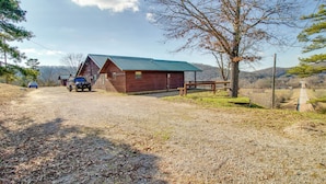 A rustic cabin with a metal roof, a dirt driveway, and a fenced area with a bench.
