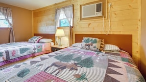 A cozy cabin-style room with two beds, a nightstand with a lamp, and a window with curtains.