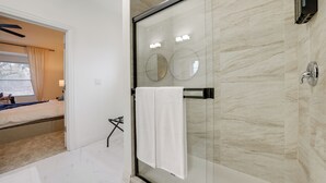 Une salle de bain moderne avec une douche à parois de verre, une serviette blanche suspendue à un porte-serviettes, et un lit visible à travers une porte ouverte.