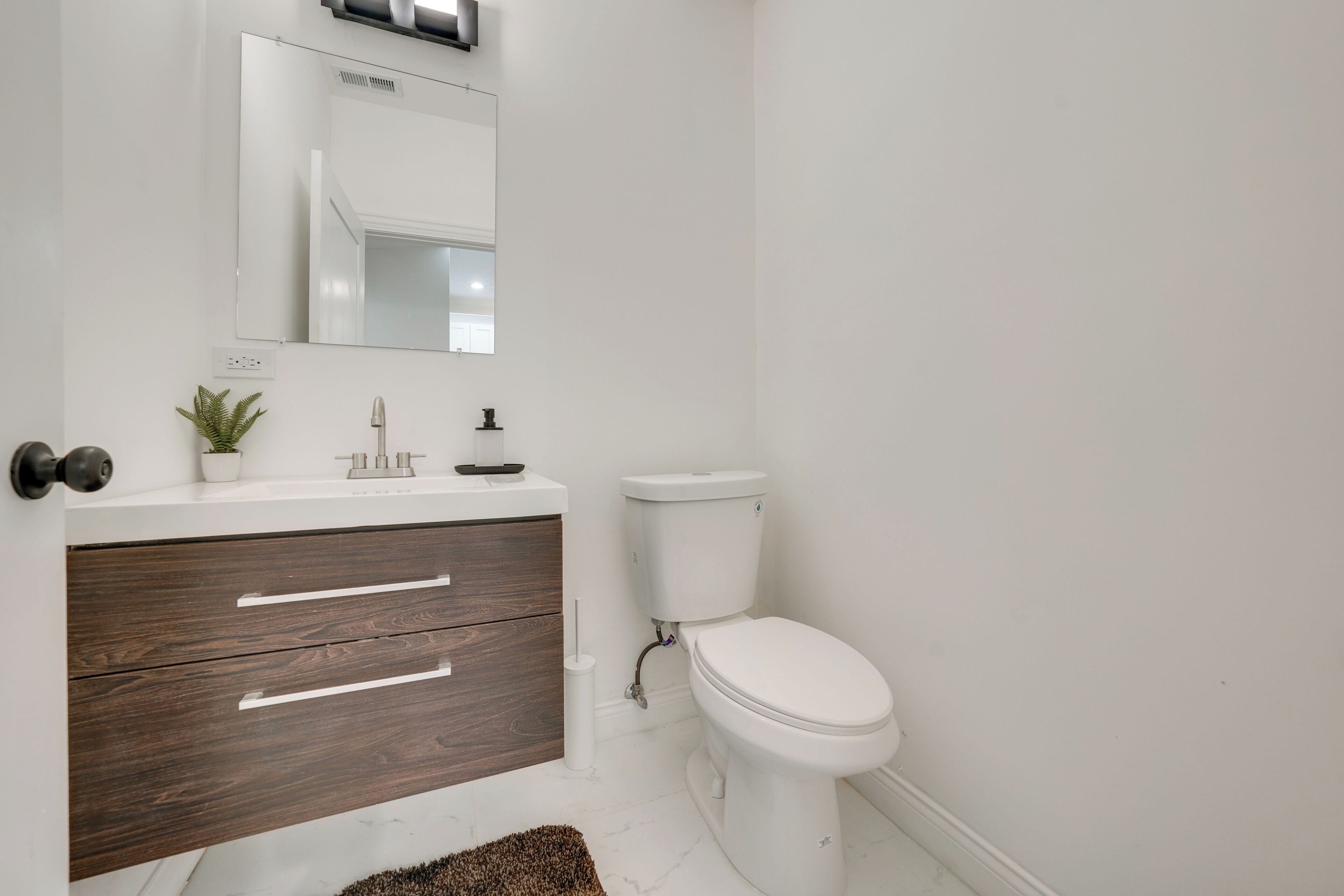 A modern bathroom with a white toilet, a wooden vanity with a mirror, and a small plant on the counter.