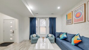 Un salon moderne avec une table basse en marbre blanc, deux fauteuils bleus, un canapé bleu agrémenté de coussins colorés et deux tableaux abstraits encadrés accrochés au mur.