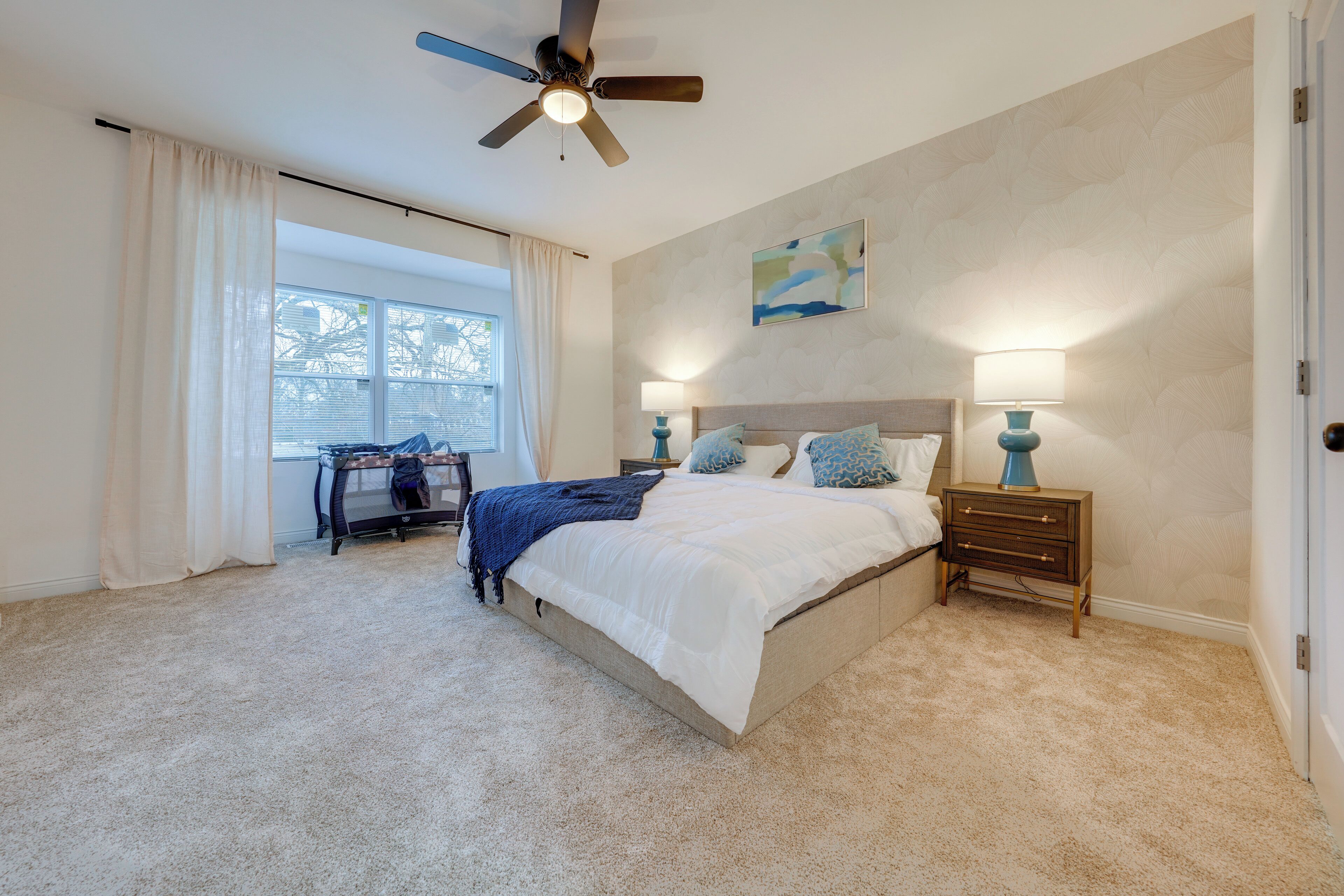 A bedroom with a bed, bedside lamps, a ceiling fan, a window with curtains, and a small table with a chair.