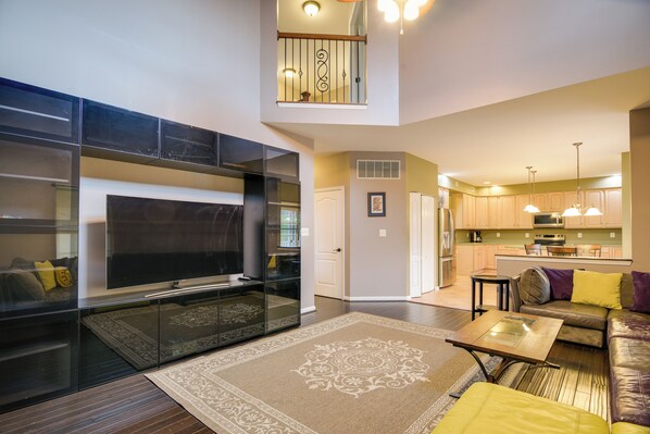 A modern living room with a flat-screen TV, a sofa, and a coffee table. There is a kitchen area with wooden cabinets and a dining table. - 54 Mi to Dtwn DC: Spacious Family Home! (Baltimore)