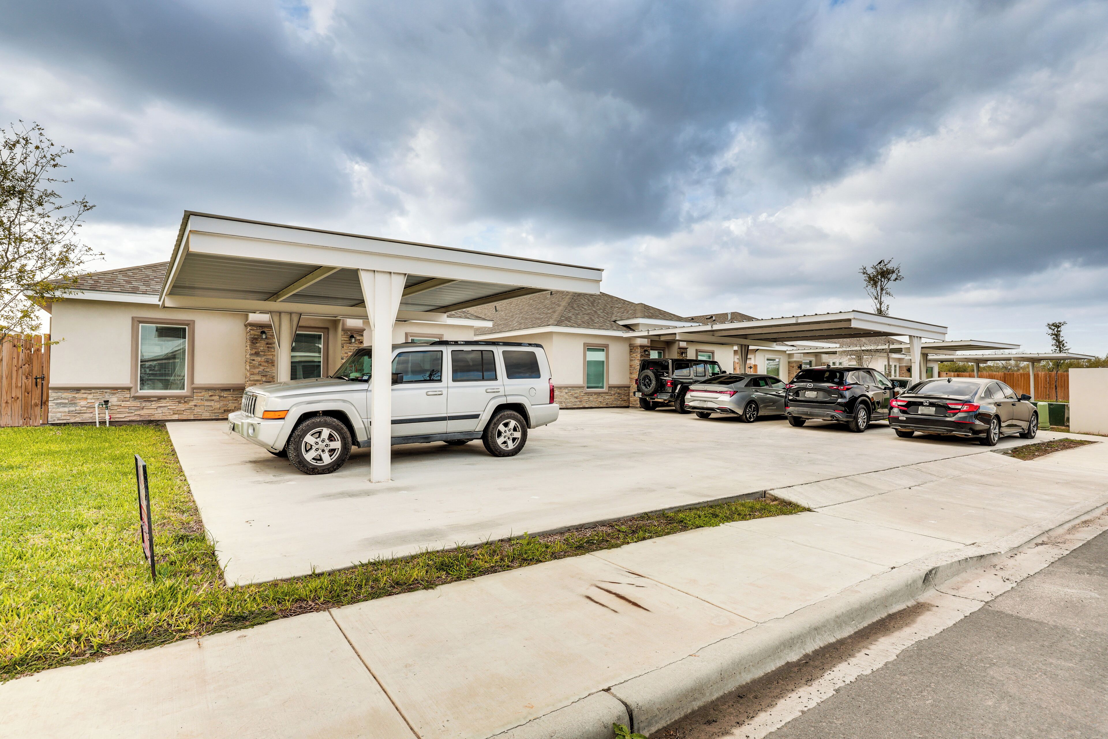 A residential property with a covered driveway, multiple parked vehicles, and a well-maintained lawn.