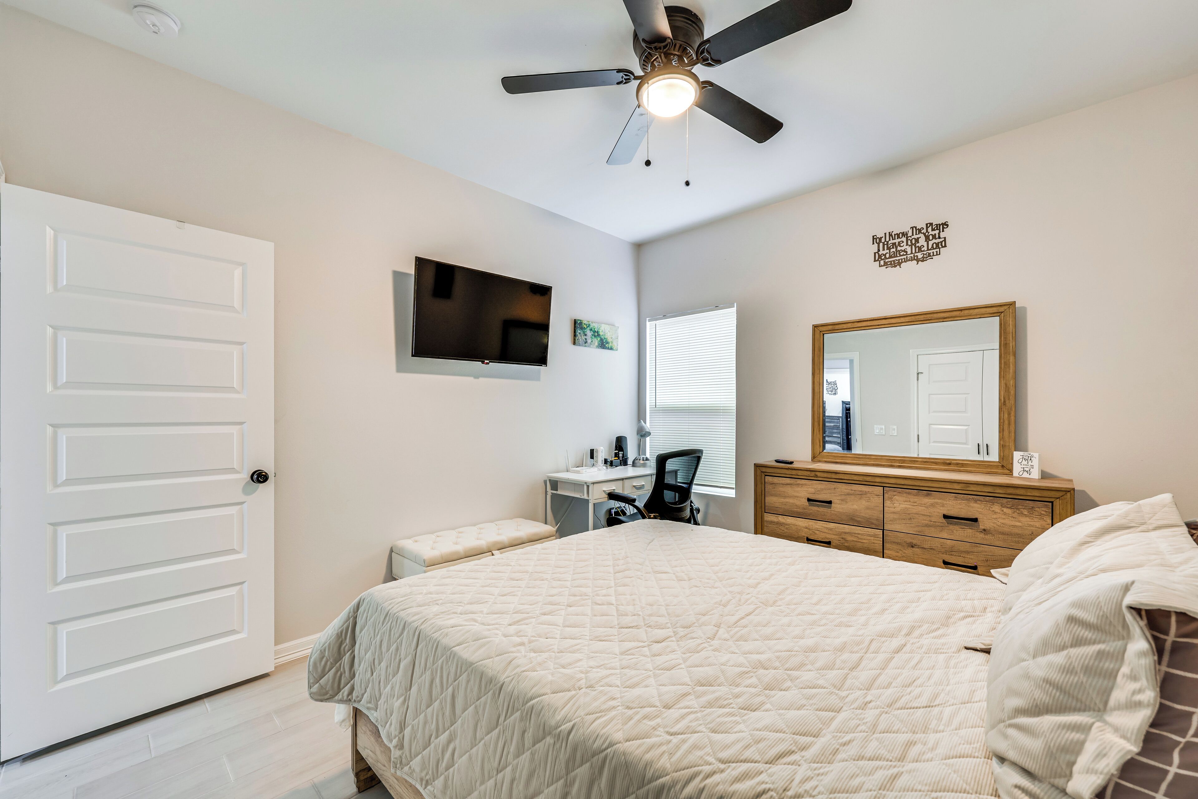 A bedroom with a bed, a desk, a chair, a dresser, a ceiling fan, a TV, and a door.
