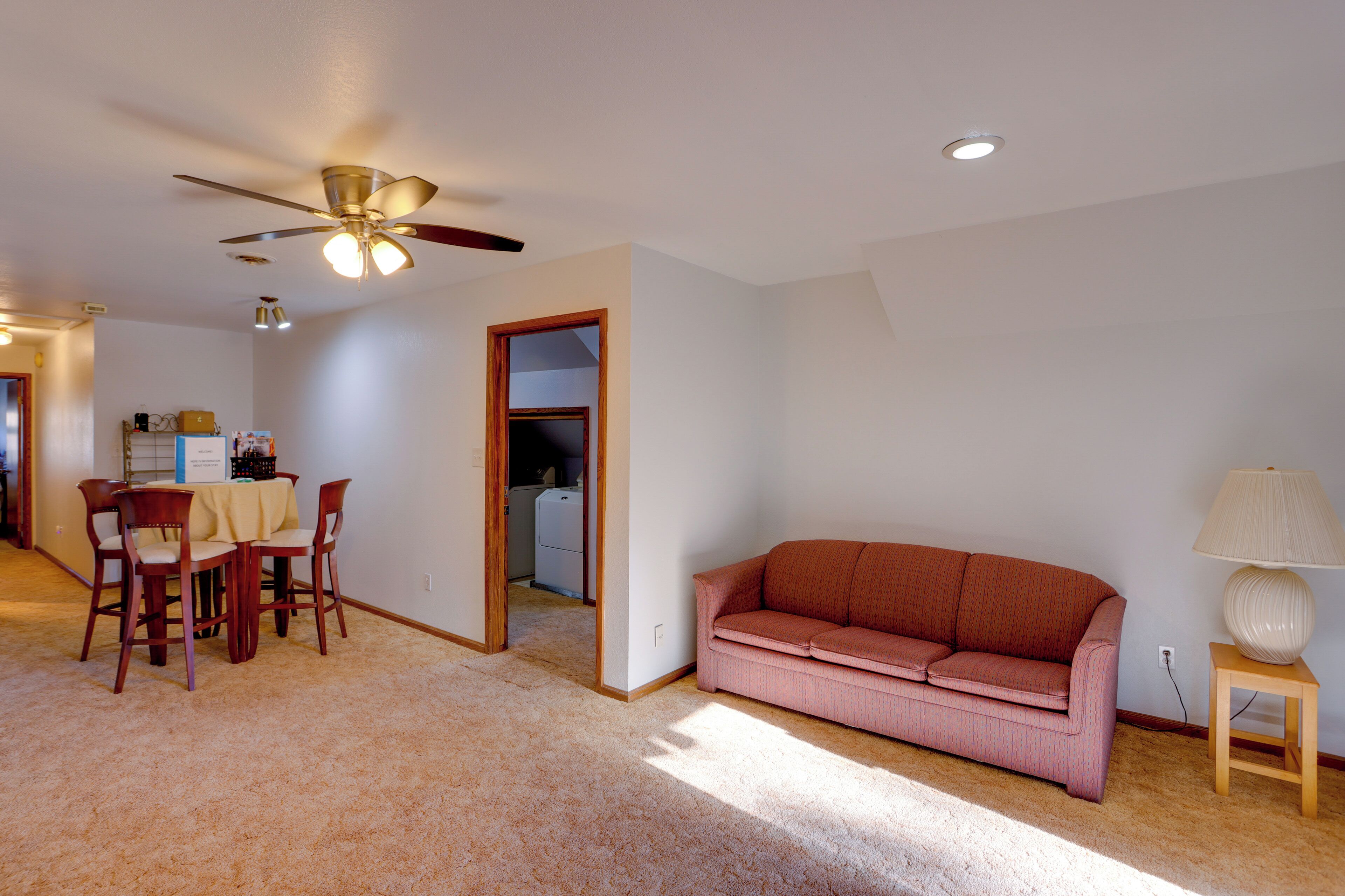 A living room with a ceiling fan, a pink sofa, a wooden side table with a lamp, and a dining area with a table and chairs.