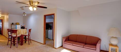 A living room with a ceiling fan, a pink sofa, a wooden side table with a lamp, and a dining area with a table and chairs.
