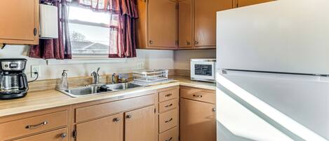 Una cuina compacta amb armaris de fusta, un refrigerador blanc, un microones, una cafetera i una finestra amb cortines.