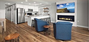 A modern kitchen with white cabinets, a stainless steel refrigerator, and a built-in microwave. There is a dining area with a wooden table and blue chairs. A fireplace with a flat-screen TV mounted above it is visible.