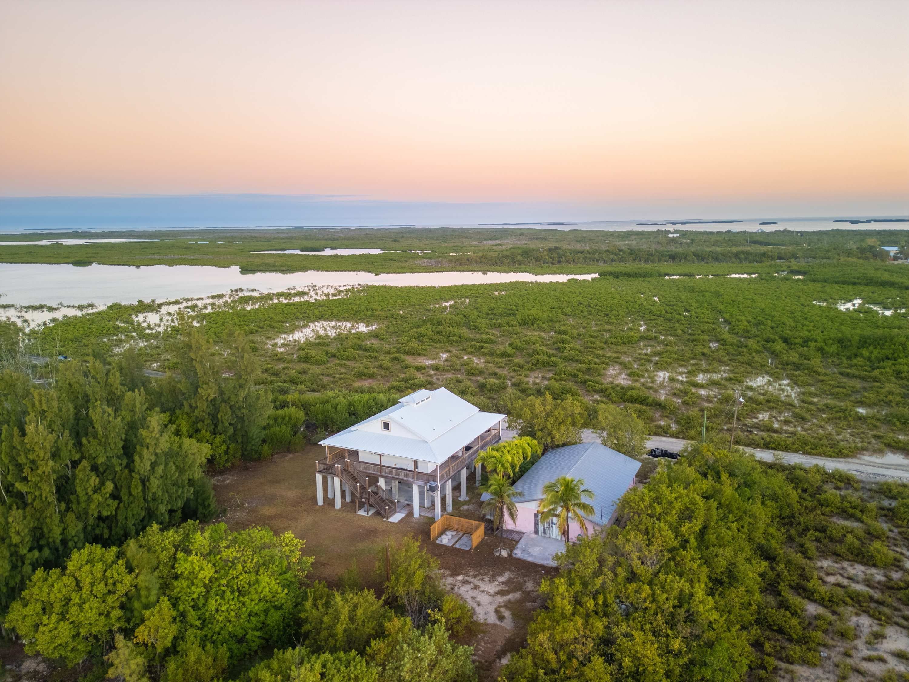 An aerial view of a large, white, two-story building with a flat roof, surrounded by dense greenery and a body of water in the background.