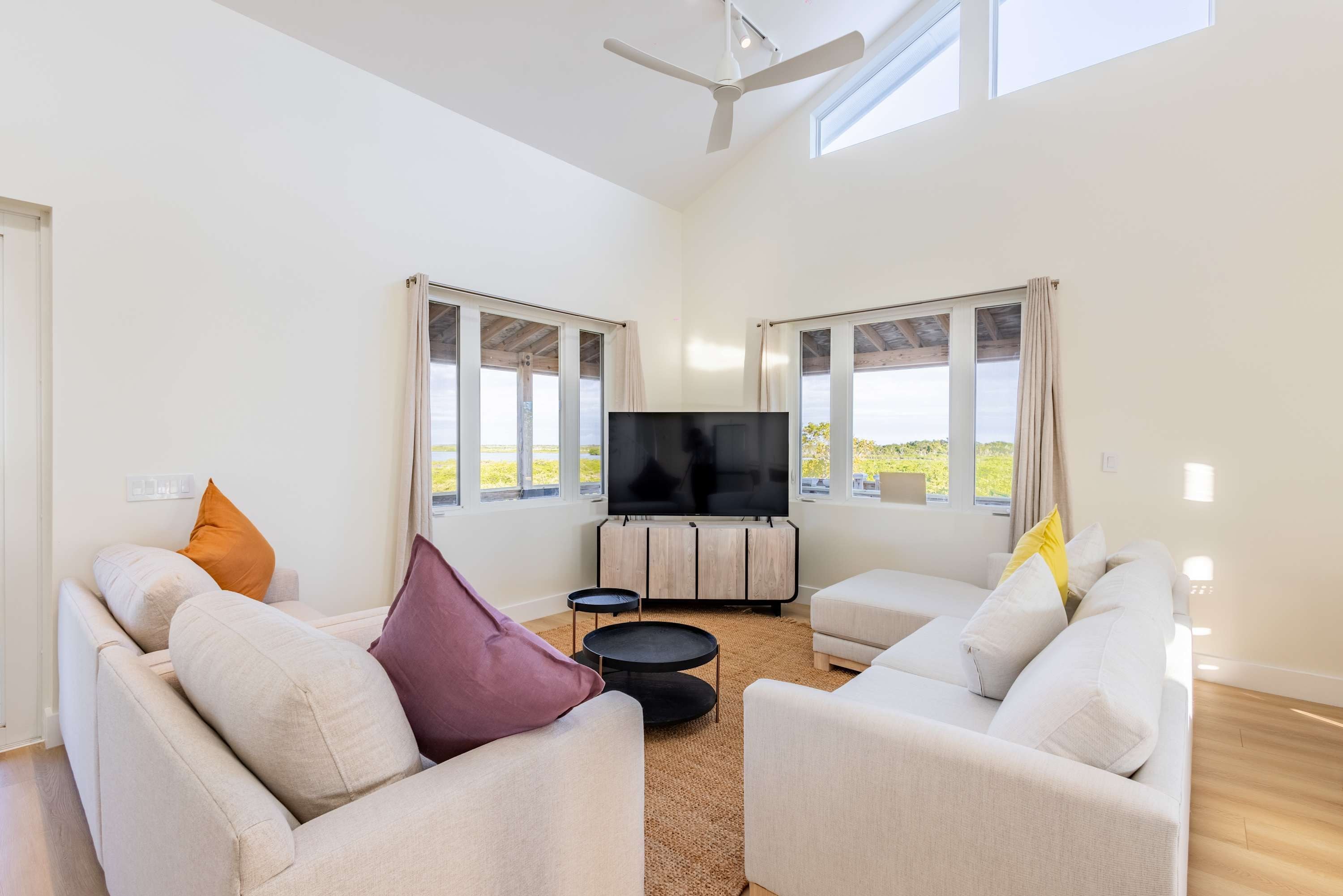 A spacious living room with a flat-screen TV, two sofas, a coffee table, and large windows offering a view of the outdoors.