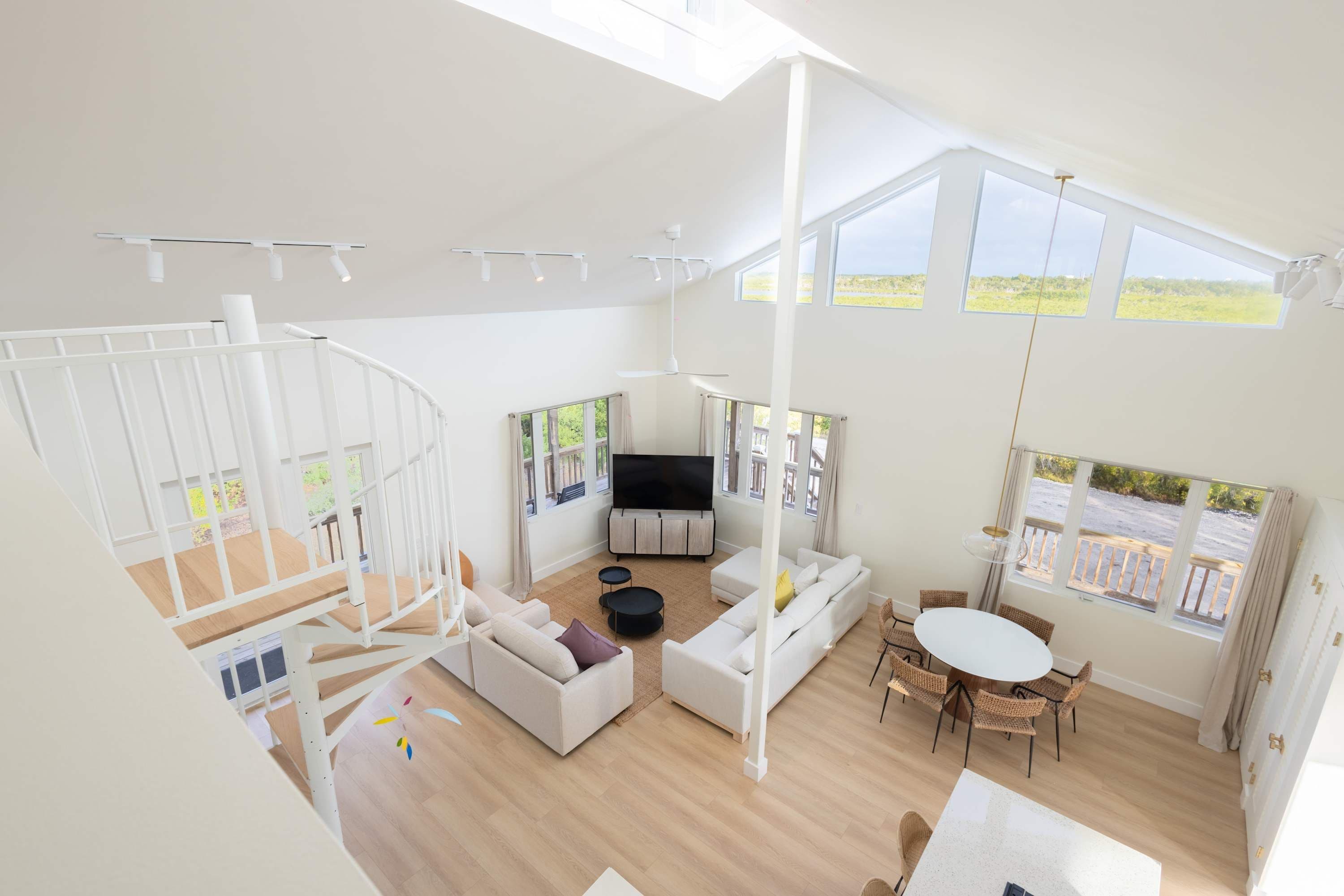 A spacious living area with a white sofa set, a TV, a dining table with chairs, and a staircase.
