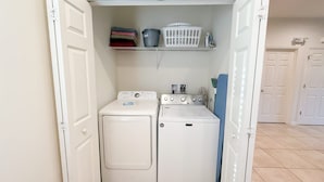 A compact laundry room with two washing machines and a shelf above containing folded clothes and laundry baskets. - Upstay Spacious 3BR Home w Resort Access (Kissimmee)