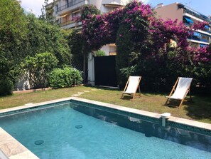 Una piscina con dos sillas de reposo vacías, rodeada de exuberante vegetación y plantas con flores.