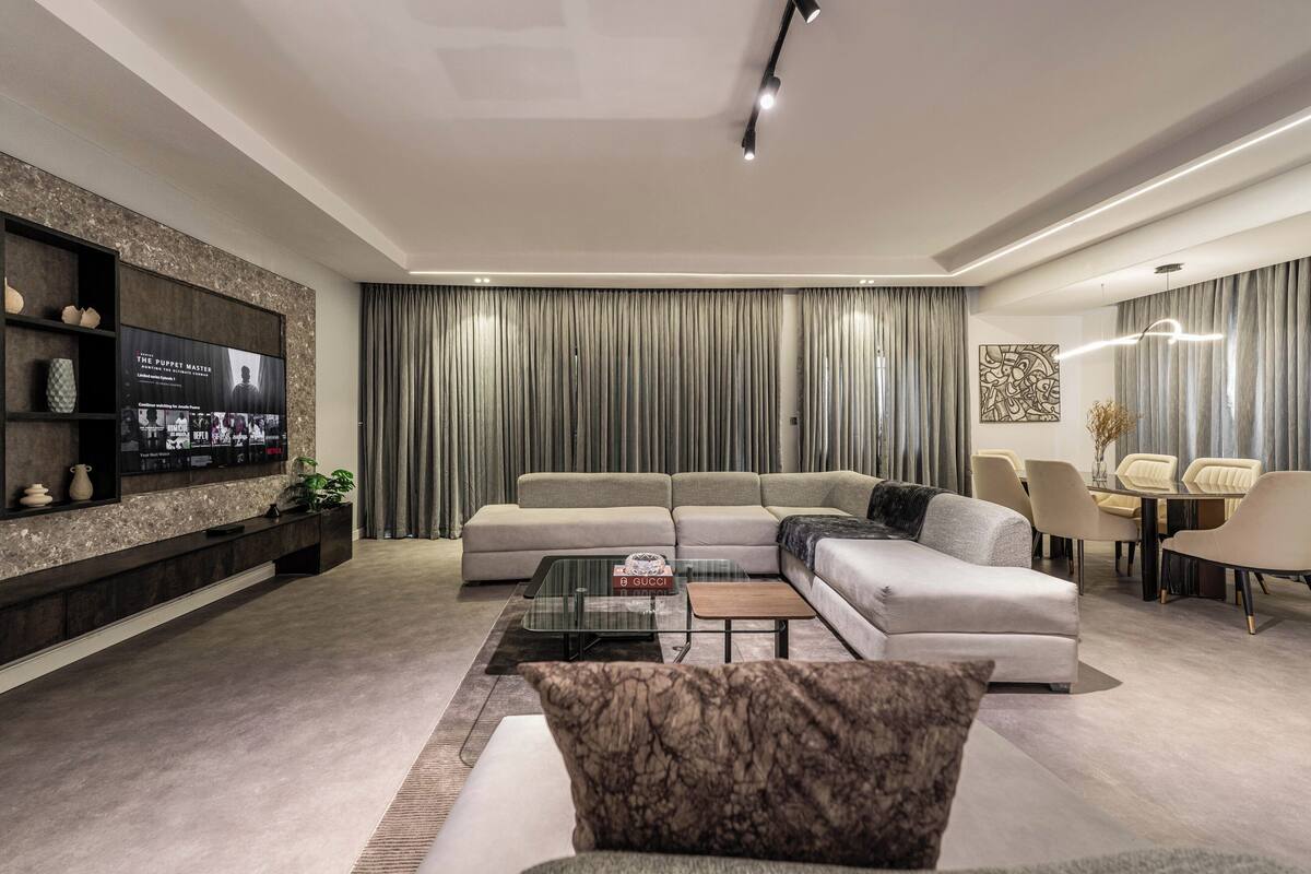 A modern living room with a sectional sofa, a glass coffee table, and a flat-screen TV mounted on a dark wall.