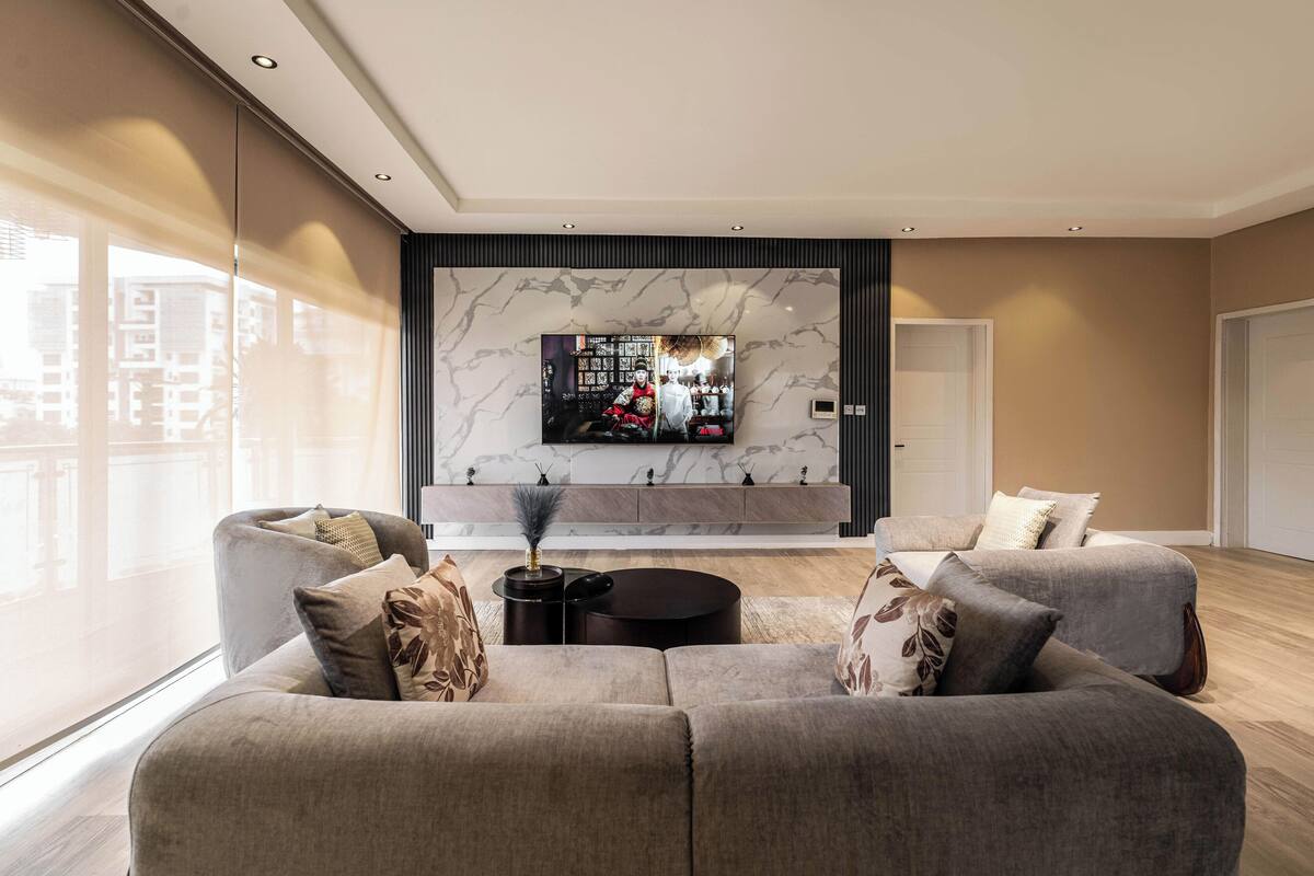 A modern living room with a large flat-screen TV mounted on a marble wall, a grey sectional sofa, a small black side table, and a large window offering a city view.