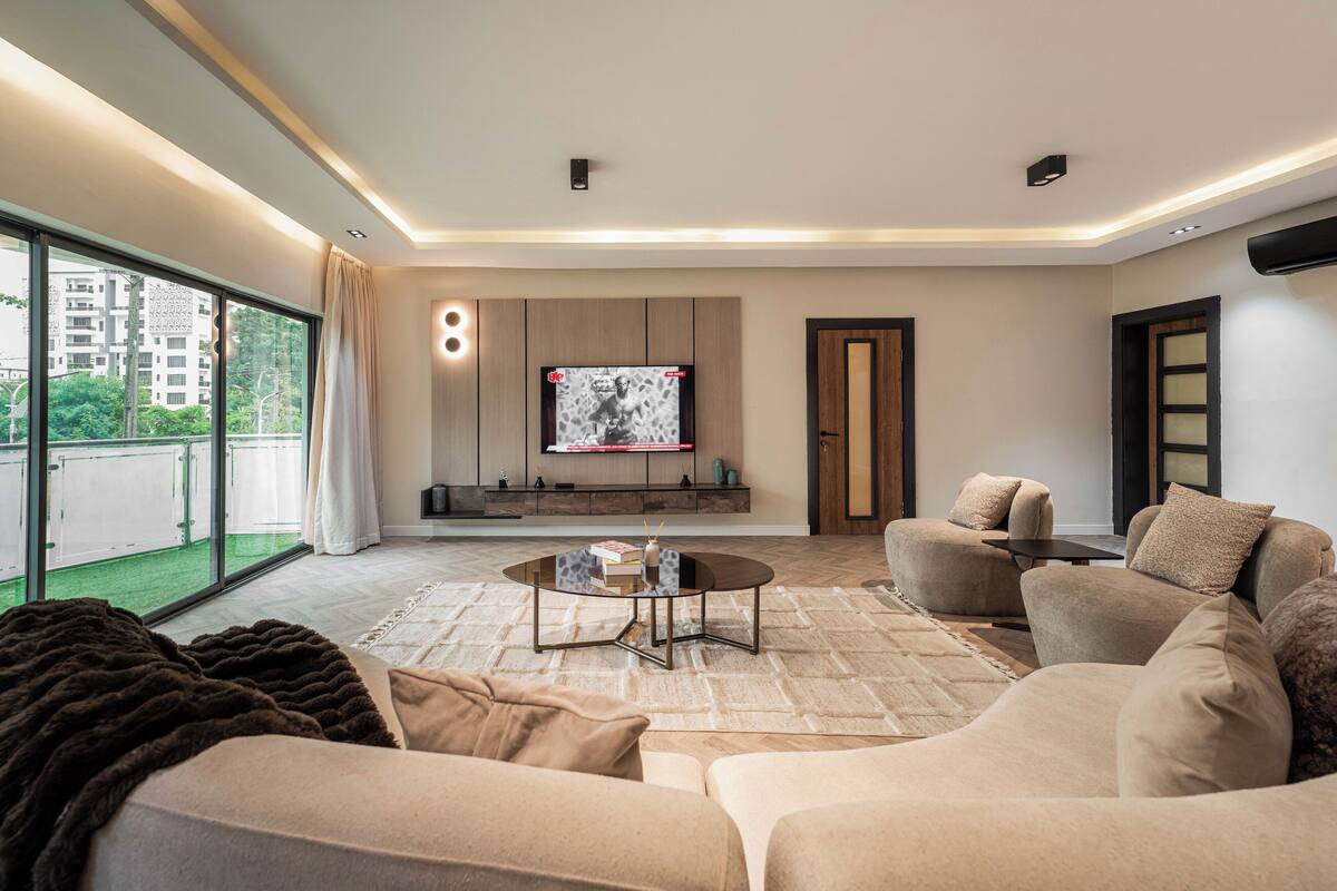 A modern living room with a large window, a flat-screen TV mounted on the wall, and a cozy seating area with sofas and a coffee table.