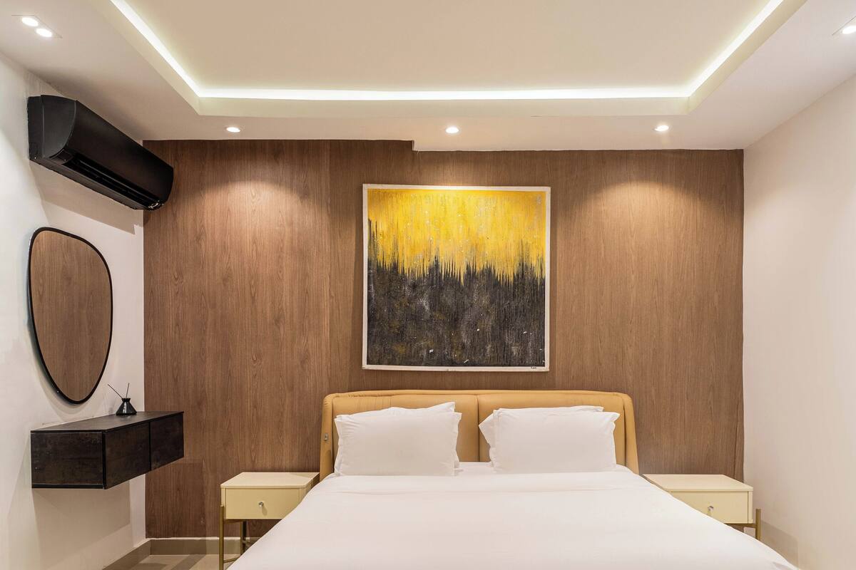 A modern bedroom with a large bed, two bedside tables, a wall-mounted mirror, and a painting.