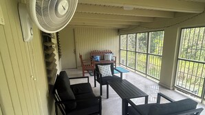 Ein überdachter Patio-Bereich mit Ventilator, Sitzgelegenheiten und großen Fenstern mit Blick auf Grünflächen.