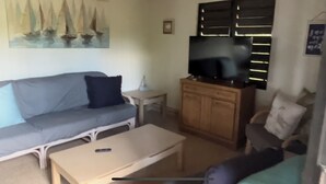 Ein Wohnzimmer mit grauem Sofa, einem kleinen weißen Tisch, einem Holzschrank und einem Fernseher darauf.