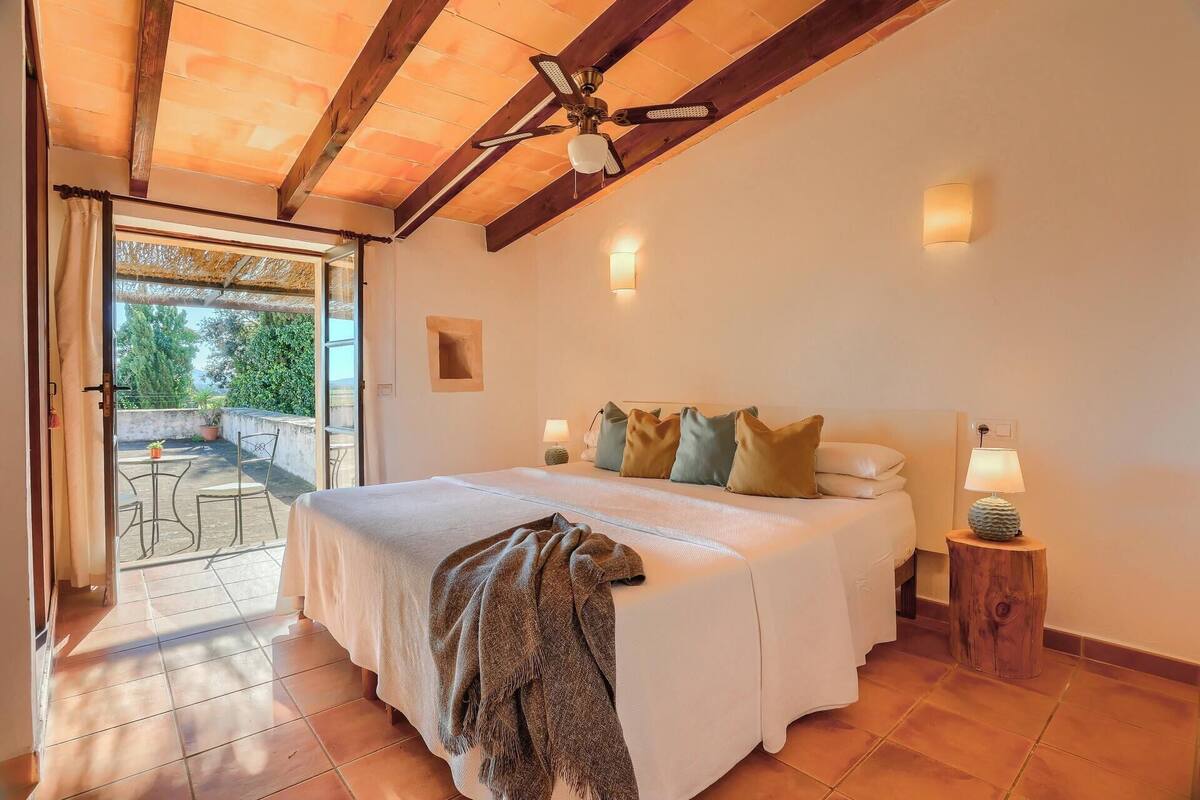 A bedroom with a large bed, a ceiling fan, a small table with a lamp, and a view of the outdoors through a glass door.