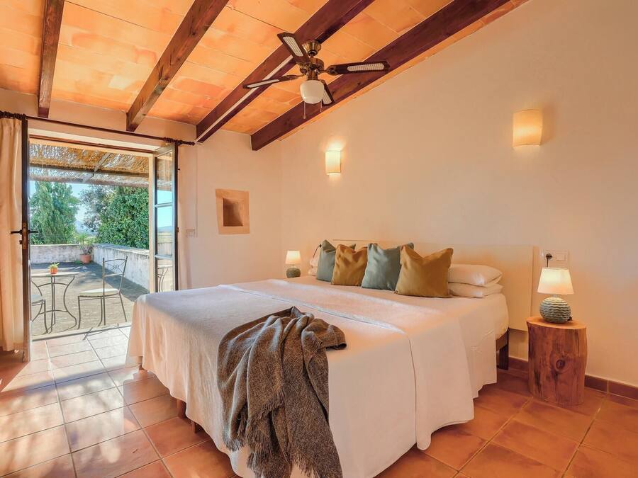 A bedroom with a large bed, a ceiling fan, a small table with a lamp, and a view of the outdoors through a glass door.