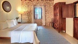 A bedroom with a stone wall, wooden beams, a bed, a nightstand, a lamp, a wardrobe, and a window. - Finca Son Valls (Felanitx)