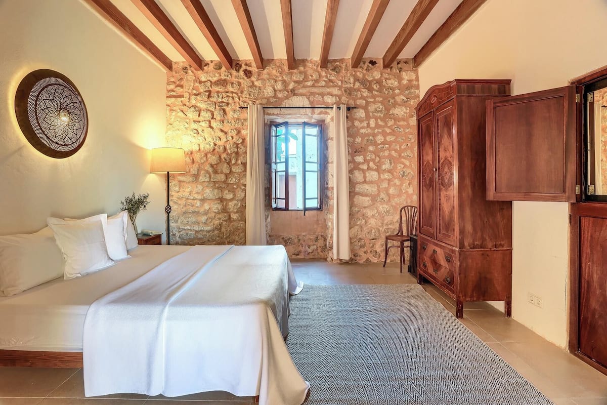 A bedroom with a stone wall, wooden beams, a bed, a nightstand, a lamp, a wardrobe, and a window.