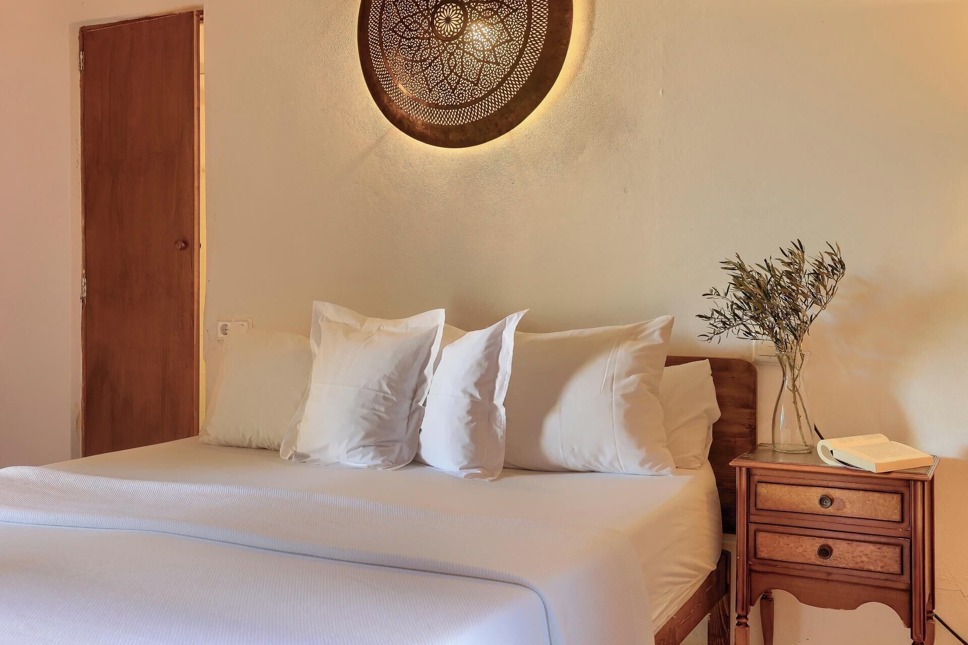 A neatly made bed with white linens, a wooden nightstand with a vase of dried flowers, and a decorative wall hanging above the bed.