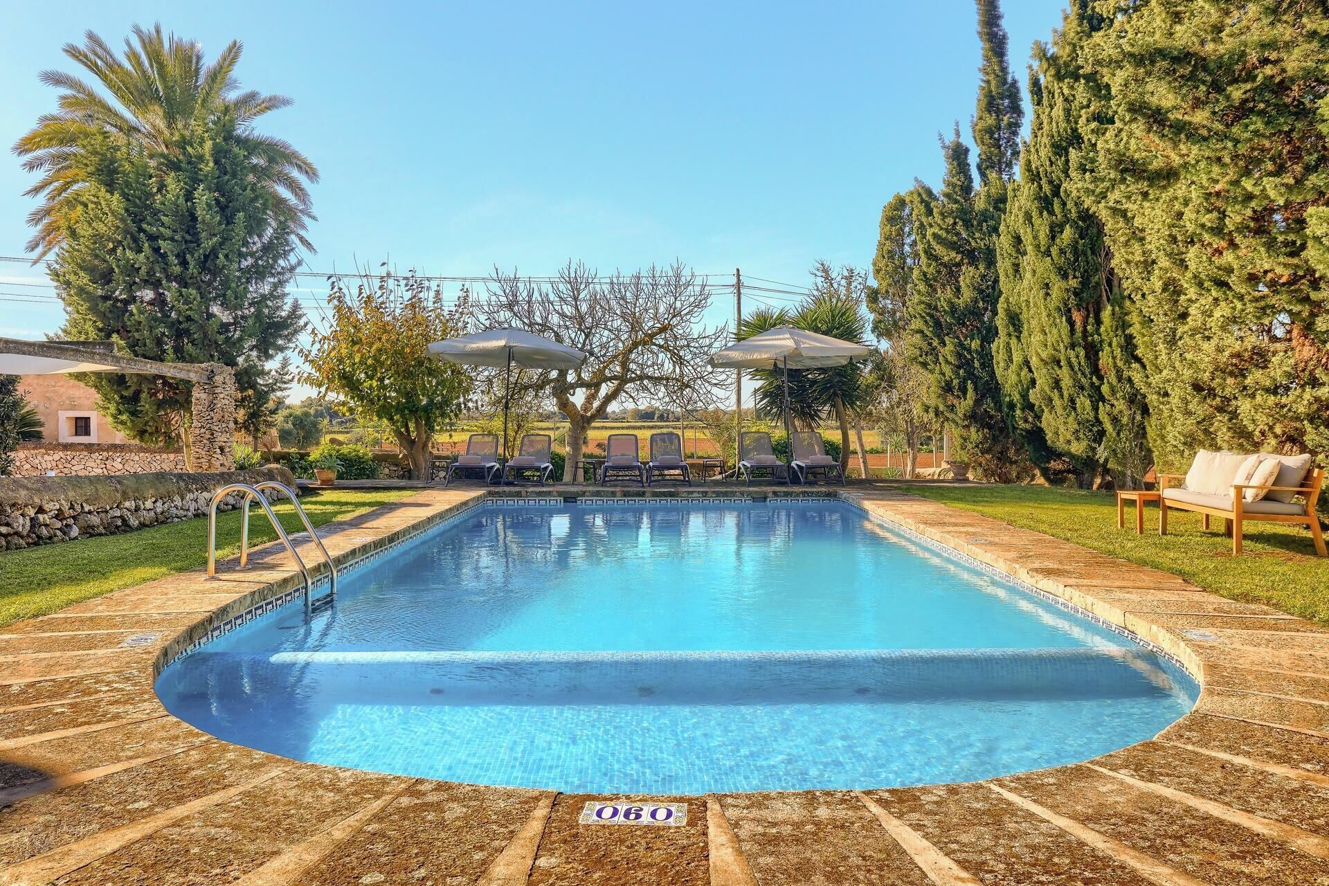 Una piscina con sillas de descanso y sombrillas, rodeada de árboles y un muro de piedra.