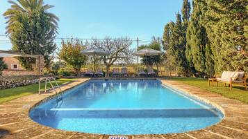 Una piscina con sillas de descanso y sombrillas, rodeada de árboles y un muro de piedra.