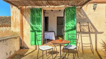 Un pequeño espacio al aire libre con una mesa y dos sillas, una escalera, y una puerta con contraventanas verdes que da a una habitación.