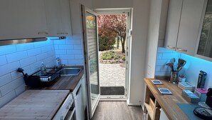 Una cocina compacta con encimera de madera, horno empotrado y un lavamanos. Una puerta da acceso a un área exterior con un sendero de piedra y árboles.