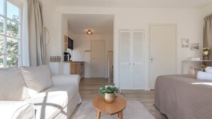 A modern, well-lit room with a sofa, a bed, a small wooden table with a plant, and a kitchen area in the background.