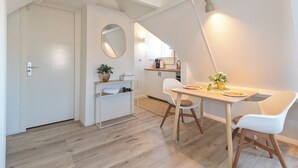 A modern, well-lit dining area with a wooden table and white chairs, a small shelf with a plant, and a mirror on the wall. - Boutique Pension Bennekom - Studio 4 (Zandvoort)