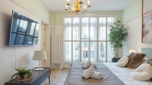 Une chambre avec un grand lit, un téléviseur à écran plat fixé au mur, une plante en pot et une fenêtre avec des stores.