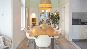 Un coin repas avec une table en bois, des chaises blanches, un luminaire suspendu et une vue sur la cuisine.