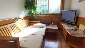 A small, cozy room with a wooden bed, a desk with a computer, and a window with a view of greenery.