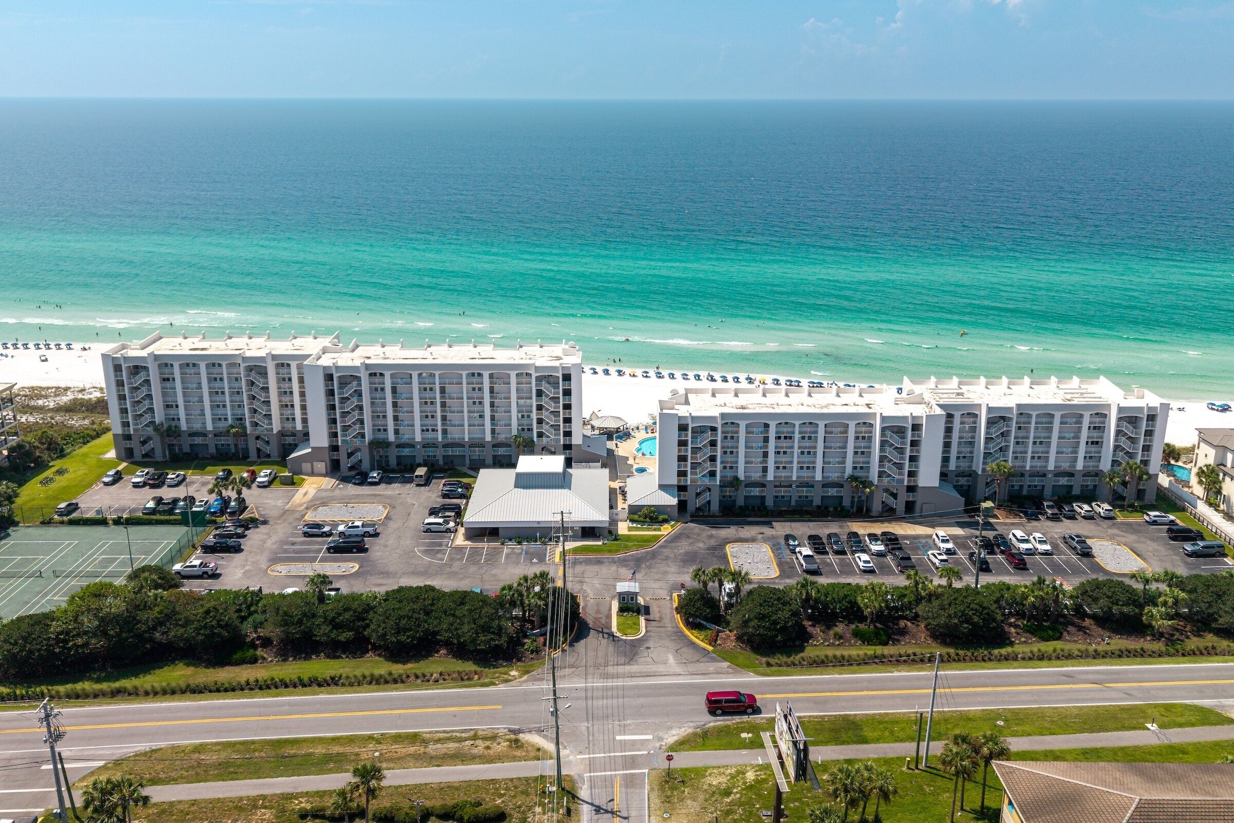 A beachfront hotel with multiple buildings, a parking lot, and a road leading to the beach.